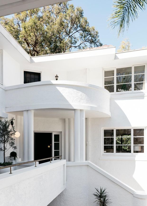 Bright white stucco residential exterior centered on a curved balcony or portico with slender round columns, black entry door, multi-pane windows, and low parapet walls. The composition shows a compact multi-level courtyard-like facade with strong sunlight, mature trees, and palms visible above the roofline.