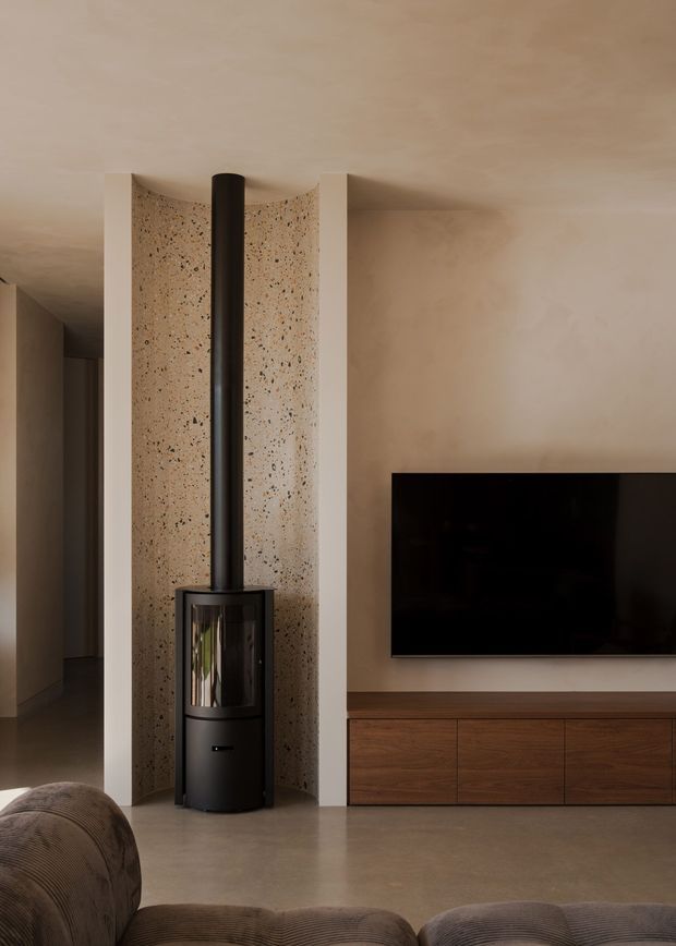 Warm, minimalist contemporary living room featuring a freestanding black wood-burning stove with a tall flue set against a curved terrazzo-look feature wall. A wall-mounted television sits above low dark timber built-in cabinetry, with neutral plaster walls, a smooth concrete-like floor, and a partial rounded upholstered sofa in the foreground.