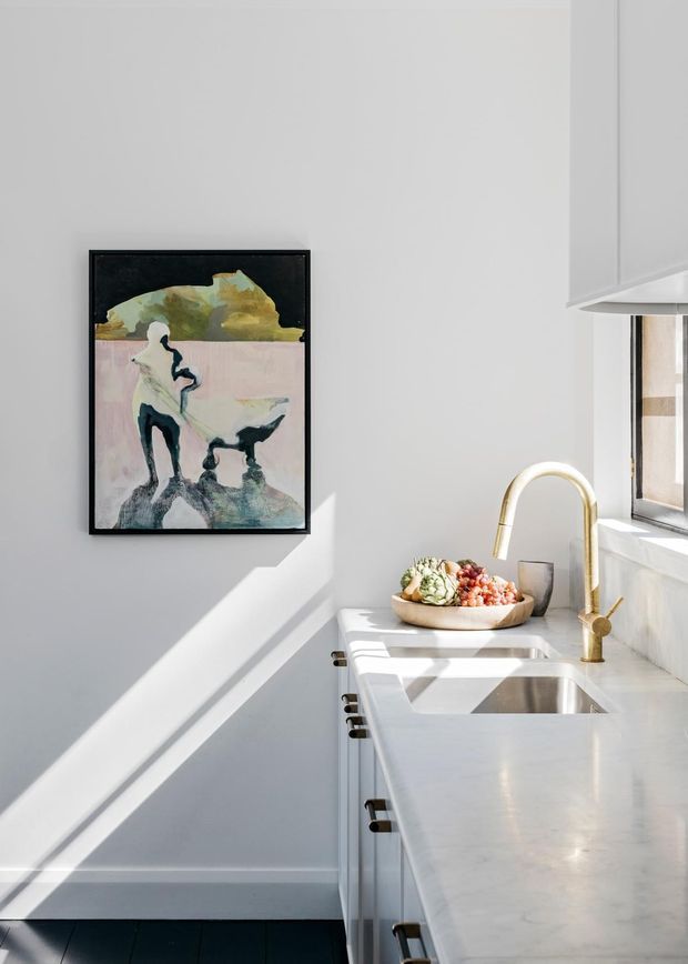 Bright, compact contemporary kitchen with white cabinetry and a pale stone countertop, featuring a deep undermount sink, sculptural brass gooseneck tap, and a sunlit window. A framed abstract artwork adds a muted organic accent to the white wall, while strong sunlight creates geometric shadows across the surfaces. Dark flooring provides contrast.