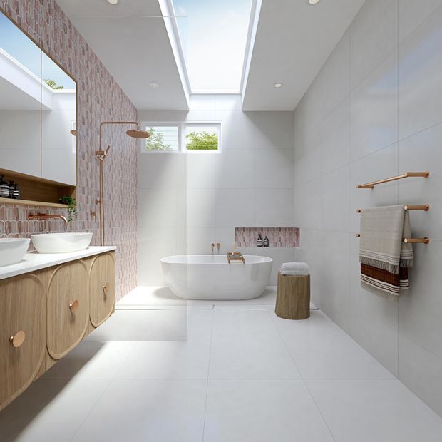 Bright contemporary bathroom with a freestanding white bathtub beneath a large skylight, a walk-in shower with warm metallic fittings, double vessel basins on curved timber cabinetry, and large pale wall and floor tiles. A patterned pink-toned feature tile wall and recessed niche add contrast, while a wooden stool and towel rail provide warm natural accents.