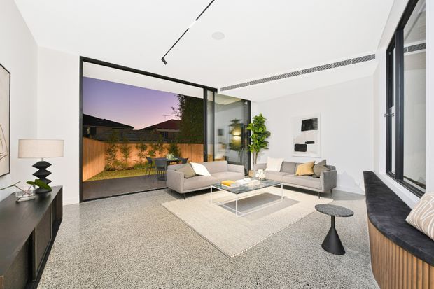 Bright contemporary living room with white walls and ceiling, polished concrete or terrazzo-like flooring, a large neutral rug, two light sofas, a glass coffee table, and minimalist black accents. Full-height black-framed glazing opens visually to a landscaped courtyard, while a built-in window seat with timber detailing runs along the right side. The room has clean lines, concealed linear lighting, and a calm indoor-outdoor connection.