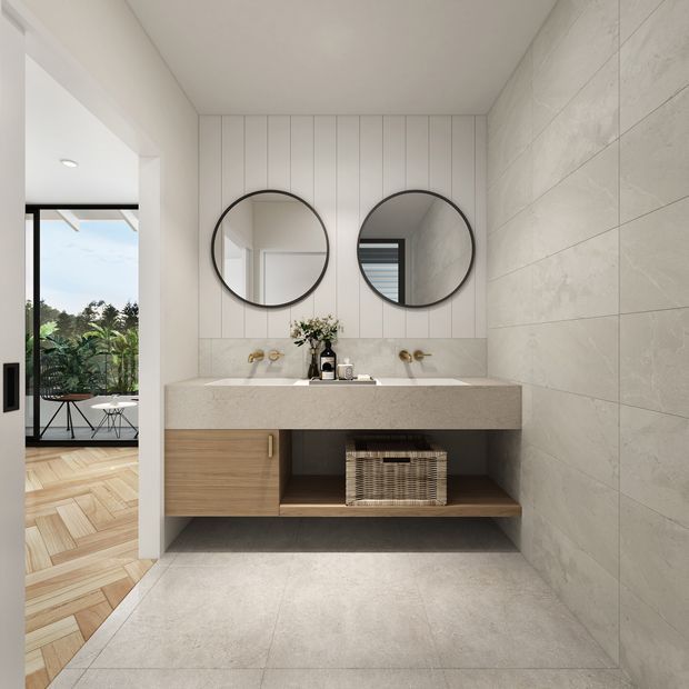 Contemporary bathroom vanity with a double basin, large round mirrors, pale stone-look tiled walls and floor, warm timber cabinetry, brass fixtures, and an open shelf. A glazed opening reveals a leafy outdoor area and adjoining timber-floored room.