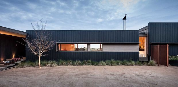 A low, contemporary residential building with a dark vertically corrugated metal-clad facade, long horizontal window band, and projecting chimney. Warm interior light glows through the glazing, while a recessed entry and timber-toned section are visible at right. A landscaped strip with ornamental grasses and a bare tree fronts the building, with a broad gravel forecourt in the foreground.