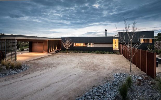 Single-storey contemporary residence viewed from a broad gravel driveway, with long low dark-clad volumes, a flat roofline, recessed warm-lit entry and carport, horizontal glazing, timber screening, and sparse native-style landscaping under an overcast sky.