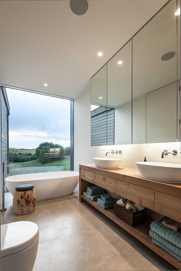 A bright, contemporary bathroom with a freestanding white bathtub beside full-height glazing overlooking a green rural landscape. The room features a long timber vanity with two white vessel basins, wall-mounted chrome fixtures, mirrored upper cabinetry, pale smooth flooring, recessed ceiling lights, and neatly arranged towels on open shelving.