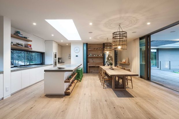 Open-plan contemporary kitchen and dining area with pale timber flooring, white cabinetry, a central island, open shelving, a long timber dining table, and large sliding glass doors opening to an outdoor deck. A large skylight and woven pendant lights create a bright, airy interior.