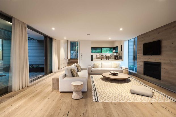 Open-plan contemporary living room with pale timber flooring, white upholstered sofas, a patterned area rug, and a round timber coffee table. Full-height glazed sliding doors with curtains open to a covered outdoor area, while a dark timber feature wall incorporates a wall-mounted television and built-in fireplace. The rear of the room opens to a compact kitchen with an island or breakfast counter.