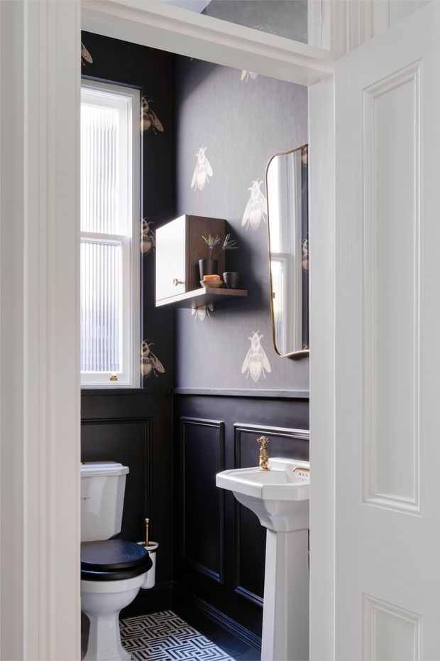 A compact, elegant powder room viewed through an open white paneled door. The room features dark charcoal lower wall panelling, grey patterned wallpaper with white insect motifs, a white pedestal basin with brass tapware, a matching white toilet, a tall window with vertical blinds, a curved wall mirror, and a small dark timber shelf with decorative objects. Geometric black-and-white floor tiles add a bold graphic accent.