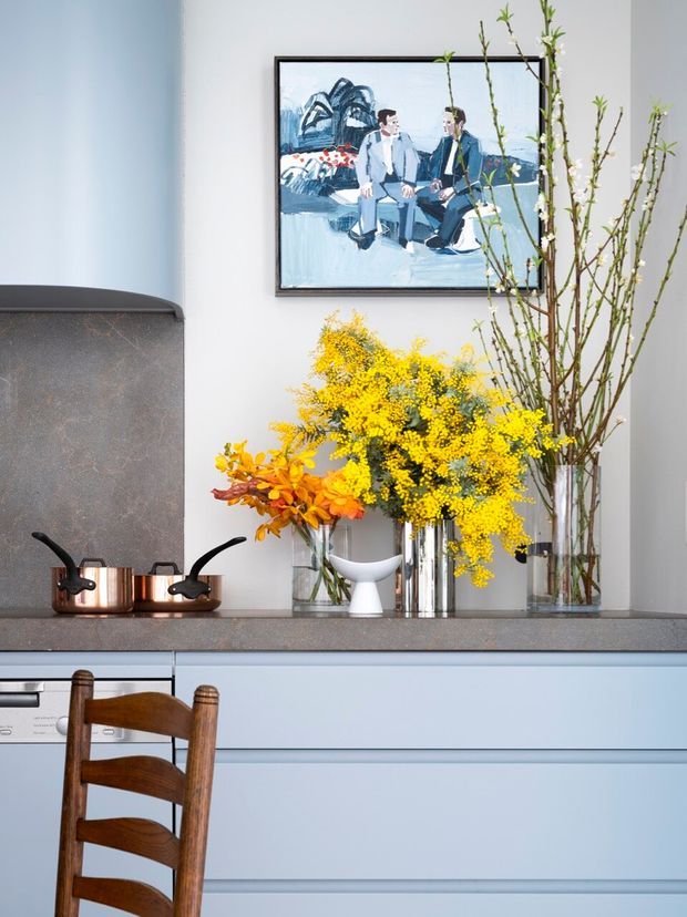 A carefully styled kitchen vignette with pale blue-grey cabinetry, a dark stone backsplash and countertop, copper cookware, and glass vases filled with bright yellow and orange branches. A framed artwork hangs above the counter, while a timber ladder-back chair is visible in the foreground. The scene has a refined, colourful contemporary-traditional character.