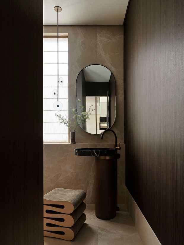 A narrow, sophisticated bathroom or powder room viewed through a dark doorway. The space features a sculptural dark stone pedestal basin with a black curved faucet, an oval wall mirror, warm taupe stone-effect wall and floor finishes, dark vertically textured wall panelling, and a tall slim pendant light beside a textured translucent window. Stacked sculptural wooden stools and a small vase with branches add understated decorative detail. The atmosphere is moody, refined, and luxurious.