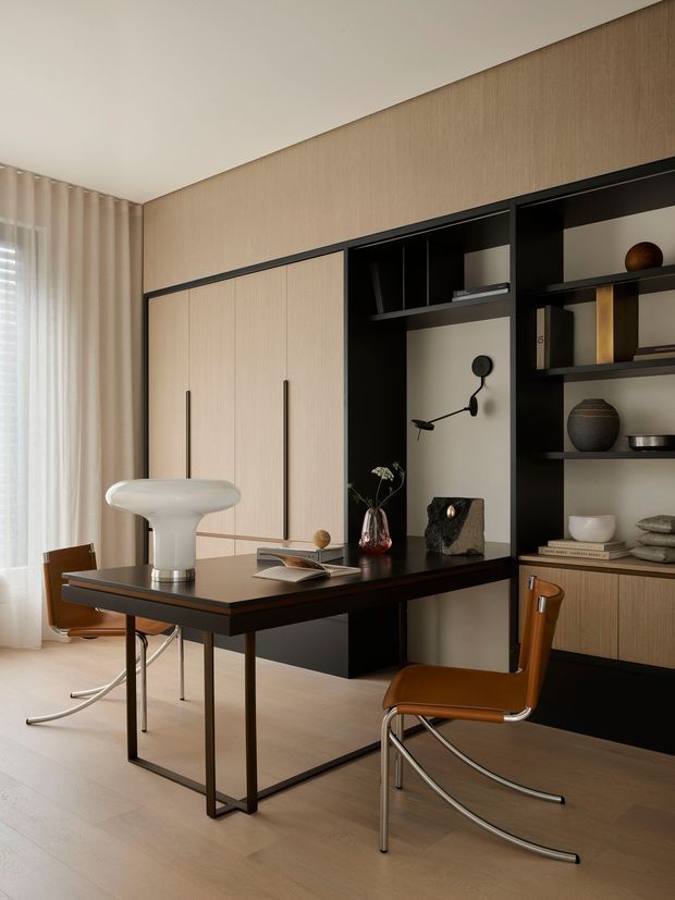 A refined contemporary dining and living area with a dark rectangular dining table, sculptural white table lamp, and two brown leather and chrome-framed chairs. Behind the table is a full-height built-in storage wall combining light timber veneer cabinetry with black open shelving and a pale display niche. Neutral textured wall covering, sheer curtains, timber flooring, curated ceramics and books, and soft natural light create a warm, sophisticated interior.