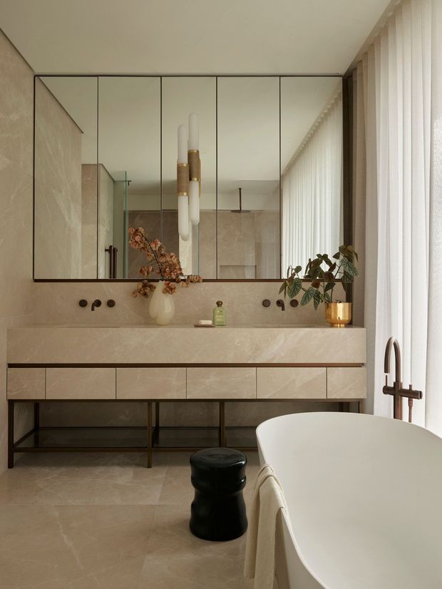 A refined contemporary bathroom featuring a freestanding oval bathtub beside a large window with sheer curtains. A long floating stone vanity with integrated basin, dark metal open support frame, and expansive mirrored cabinet spans the rear wall. Warm beige stone surfaces, black fixtures, sculptural pendant lighting, and restrained botanical accents create a calm, luxurious atmosphere.