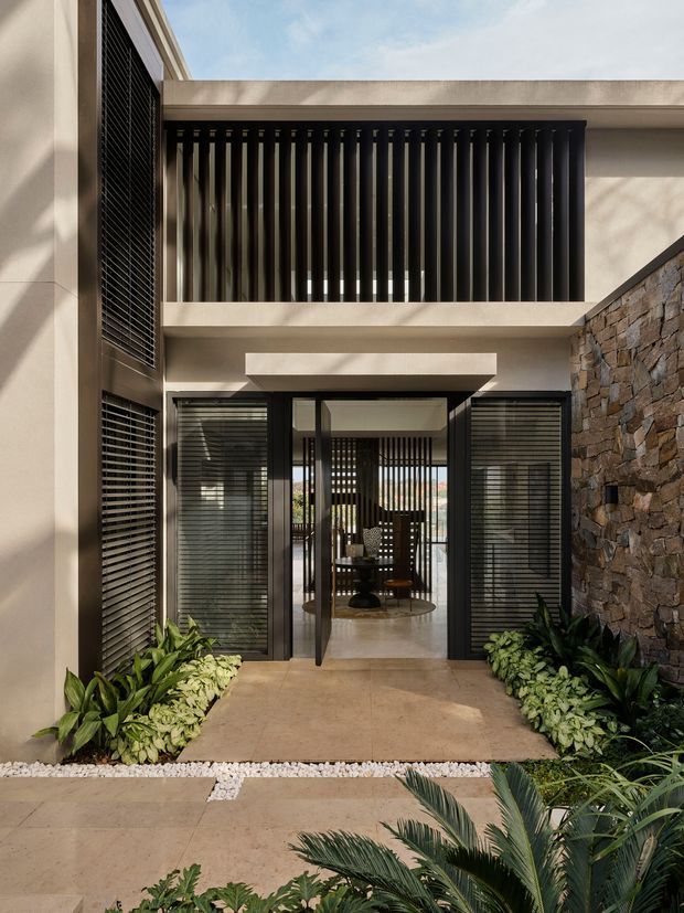 A refined modern residential entry courtyard viewed from outdoors. The two-storey facade combines smooth warm beige rendered walls, extensive dark vertical and horizontal louvres, glazed entry doors, and a rough stacked-stone feature wall. Low tropical planting borders the concrete-paved approach, with a bright open sky above.