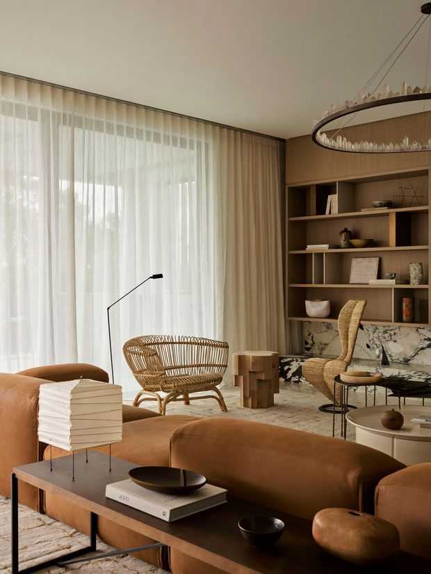 A warm, refined living room with floor-to-ceiling sheer curtains, a large brown leather sectional sofa, a woven rattan lounge chair, and a dark wood coffee table. The room features built-in timber shelving with marble-backed cabinetry, sculptural decor, a linear pendant light, and neutral textured finishes.