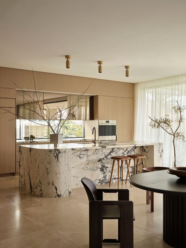 A refined open-plan kitchen and dining area with a large veined marble island, integrated timber cabinetry, built-in oven, dark sculptural tapware, timber bar stools, and a round dark dining table. Floor-to-ceiling sheer curtains bring in soft daylight, while a large interior opening and mirrored or glazed surfaces visually extend the space. The palette is warm and neutral with natural branches used as understated decoration.