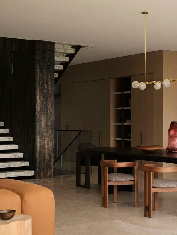 A sophisticated open-plan interior combining a dining area, living area, and stairway. The space features dark timber-toned built-in cabinetry, a dramatic black and textured timber stair wall, pale stone-look flooring, a black dining table with timber chairs, a warm tan sofa, and a sculptural brass pendant light. The overall atmosphere is refined, warm, and moody with contemporary detailing.