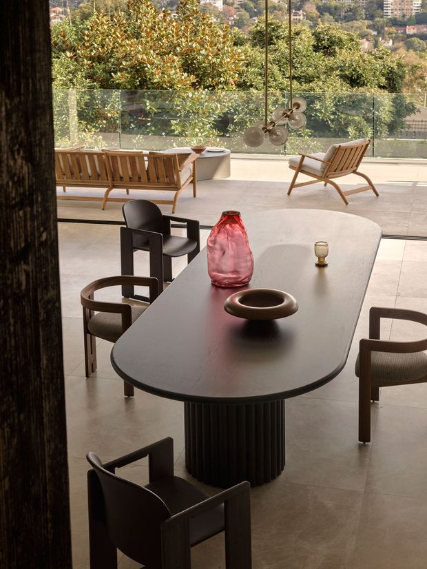 A refined dining area with a large dark oval dining table and sculptural fluted pedestal, surrounded by contemporary upholstered and timber-framed chairs. Warm decorative objects sit on the tabletop, while a glazed wall opens to a spacious terrace with timber lounge furniture and landscaped hillside views. The room has a calm, gallery-like atmosphere with abundant natural light and expansive glazing.