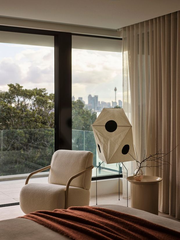 A softly styled bedroom corner with a cream upholstered armchair beside expansive floor-to-ceiling glazing. A sculptural geometric floor lamp, cylindrical side table, sheer and full-height curtains, and a rust-brown throw create a warm, refined interior. Trees and a distant urban skyline are visible beyond the glass balcony railing.