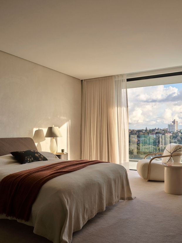 A warm, refined bedroom with a low-profile upholstered bed dressed in cream bedding and a dark rust-brown throw. Soft beige walls, floor-to-ceiling curtains, plush carpeting, sculptural bedside lamps, and a curved lounge chair create a calm, luxurious atmosphere. Full-height glazing opens to a balcony or terrace with a distant urban view.