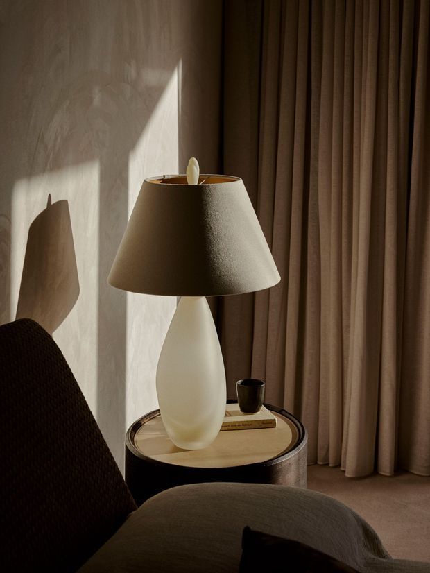 A moody, close-up interior vignette featuring a sculptural white table lamp with a textured taupe fabric shade on a round side table. The table holds a book and a small dark cup, while floor-to-ceiling taupe curtains and a softly textured neutral wall form the backdrop. A dark upholstered chair occupies the foreground, with strong sunlight creating dramatic shadows.