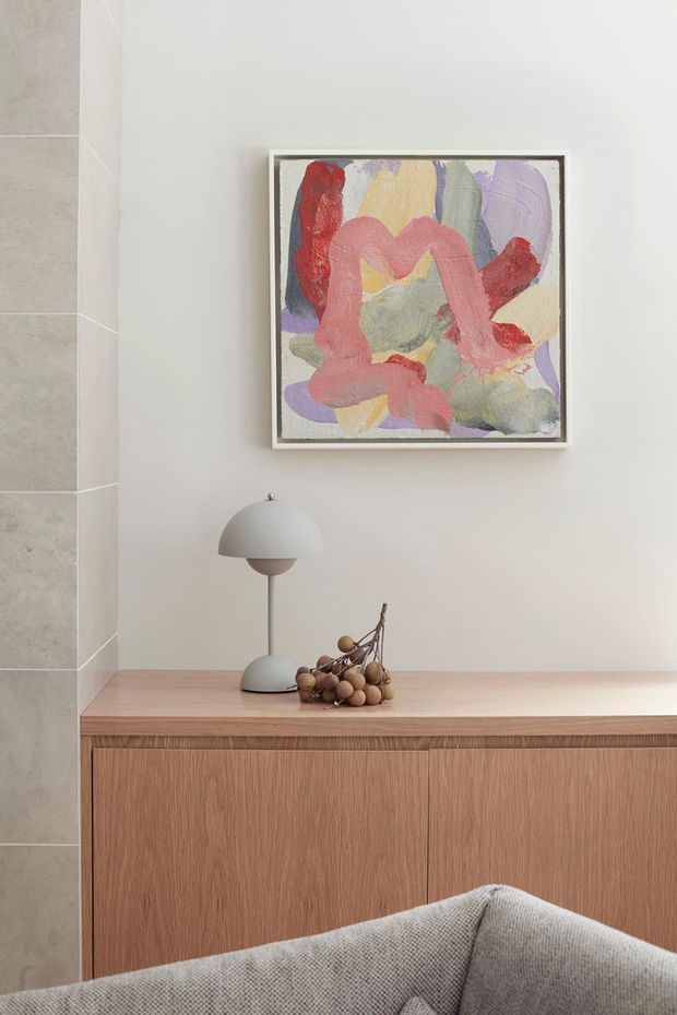 A softly styled interior vignette featuring a low timber sideboard with flush vertical-grain cabinetry, a sculptural dome-shaped table lamp, decorative branch arrangement, and colourful abstract artwork above. The neutral plaster wall and large-format grey stone-look tiled wall create a calm, refined setting, with the corner of a textured upholstered sofa in the foreground.