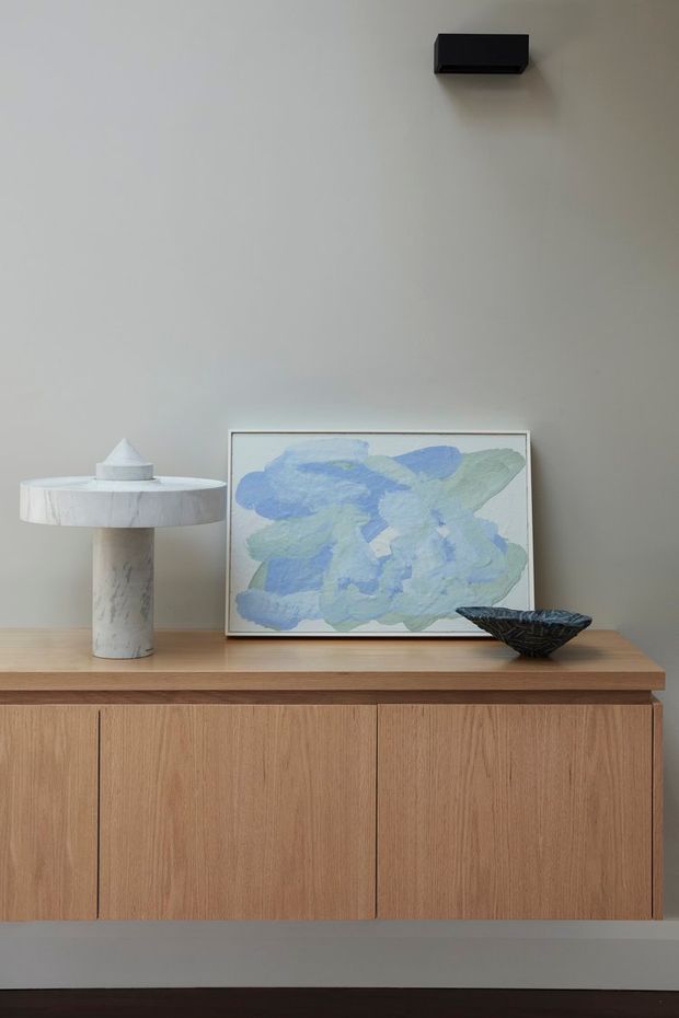 A minimalist interior vignette featuring a built-in light timber sideboard with vertical-grain cabinet doors, topped with a pale stone or timber surface. A sculptural marble-toned table lamp, abstract blue-green artwork, and dark sculptural bowl sit against a warm grey wall. A small black wall-mounted light fixture provides a subtle architectural detail.