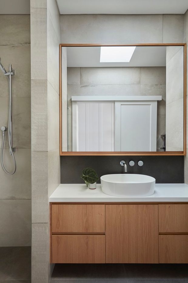 Modern bathroom vanity with a round white vessel basin, wall-mounted timber cabinetry, white benchtop, large timber-framed mirror, and a tiled shower area. Neutral grey-beige wall tiles and a dark small-format tile backsplash create a clean, contemporary fit-out. A skylight and mirrored reflection add brightness and depth.