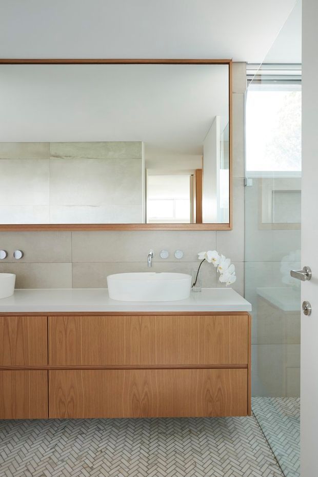 Bright contemporary bathroom featuring a floating timber vanity with a white countertop and vessel basin, large frameless mirror with timber trim, pale concrete-look wall tiles, and a glass-enclosed shower. The floor has a small-format light tile laid in a herringbone pattern, with brushed metal fixtures and a white orchid adding a subtle decorative accent.