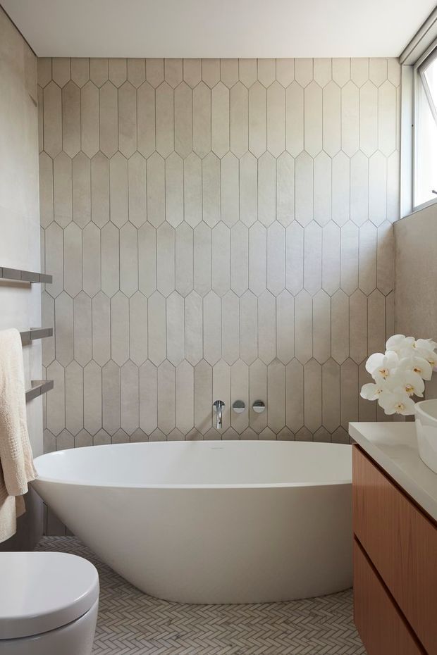 A bright, refined bathroom featuring a freestanding white bathtub, a large geometric elongated-hexagon tiled feature wall, patterned light floor tiles, a timber vanity, wall-mounted fixtures, a towel rail, and a high window. White orchids add a small decorative accent.