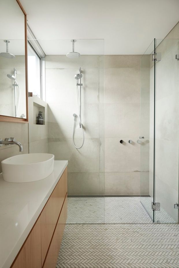 Bright contemporary bathroom with a large walk-in glass shower, dual shower heads, pale stone-look wall tiles, and a white herringbone mosaic floor. A floating timber vanity with a white vessel basin sits beneath a large timber-framed mirror, creating a clean, warm and refined interior.