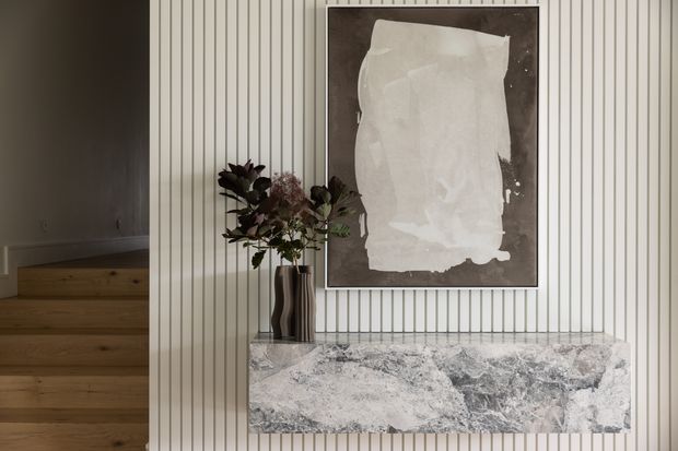 A refined interior feature wall with vertical fluted panelling, a dramatic veined stone mantel or console surface, framed abstract artwork, and a sculptural vase with foliage. Timber stairs are visible to the left, adding warmth to the otherwise neutral, gallery-like composition.