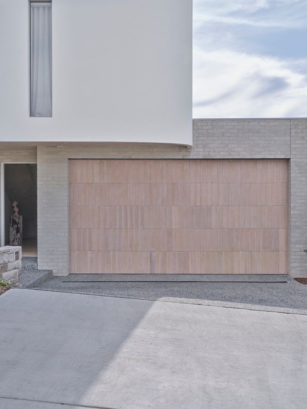 Minimalist modern residential facade viewed from the driveway, featuring a large pale timber garage door set within a light grey brick wall, smooth white rendered upper wall, narrow vertical window, and a recessed entry at the left. The composition is clean and restrained with subtle warm timber against cool neutral masonry and concrete paving.