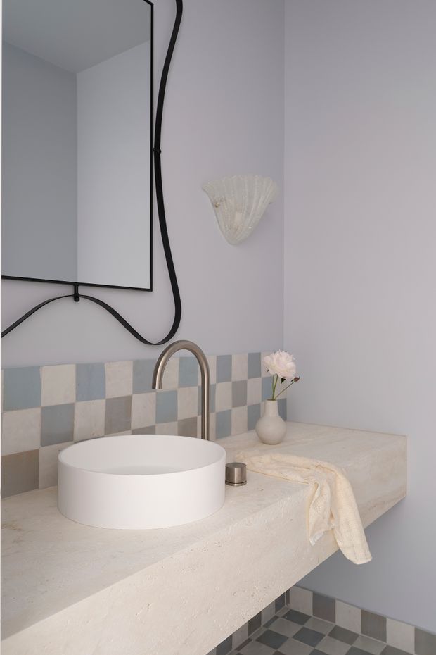 A refined contemporary bathroom vanity featuring a round white vessel basin on a thick pale stone or concrete-look benchtop, with a brushed metal gooseneck tap. The walls are painted in a soft lavender-grey and accented by a checkerboard tiled backsplash and matching floor in muted blue-grey and off-white. A large frameless or black-edged mirror, sculptural black cord, decorative wall sconce, vase, and draped towel add character to the compact space.