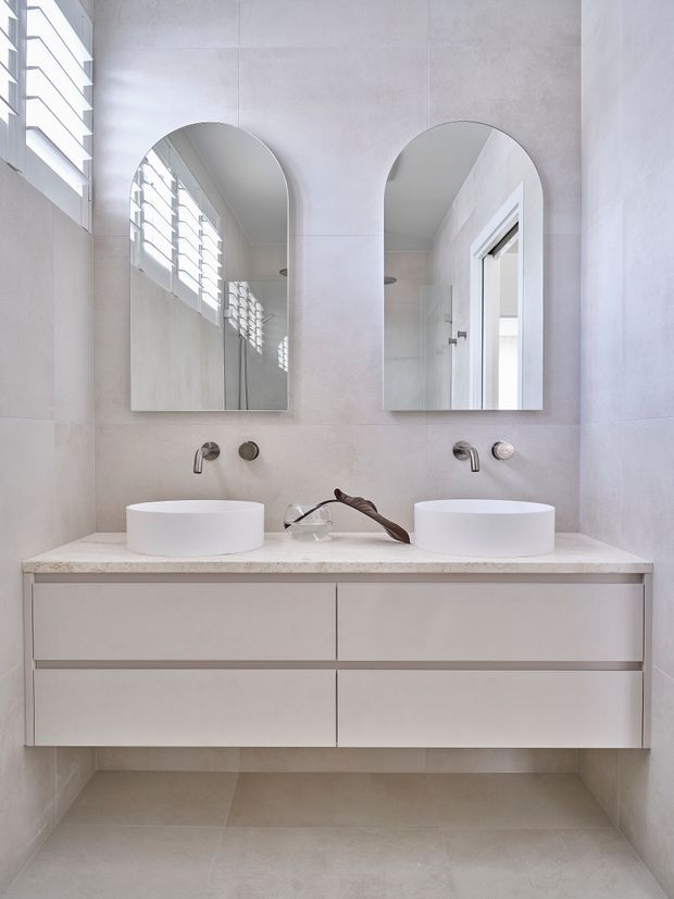 Bright, minimalist bathroom vanity with two round vessel basins, wall-mounted brushed metal taps, a pale stone benchtop, and floating white drawer cabinetry. Two arched mirrors sit above the vanity, while louvred windows provide filtered natural light. Pale large-format tiled walls and floor create a clean, understated interior.