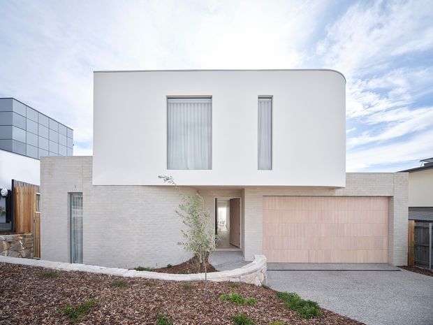 Front exterior view of a contemporary two-storey residence with a curved white upper volume, narrow vertical windows, pale brick lower facade, and an integrated double garage. A recessed entry and curved low retaining wall define the landscaped frontage.