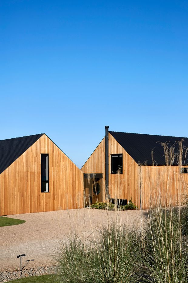 Exterior view of a contemporary timber-clad building composed of connected steep gabled volumes. The facade has vertical natural timber boards, dark corrugated roofing, narrow black-framed windows, and a gravel forecourt with ornamental grasses and low landscape lighting under a clear blue sky.