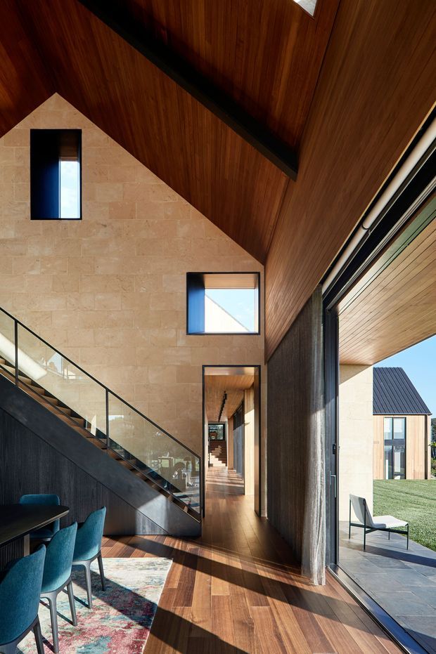 Contemporary interior view of a tall, light-filled circulation and living space with a dramatic timber-lined sloping ceiling, warm timber flooring, and a pale stone feature wall. A glass-balustraded staircase rises along the wall, beside a dining area with teal upholstered chairs and a patterned rug. Full-height glazing opens to a paved outdoor area and lawn, while smaller high-set windows bring in additional daylight.