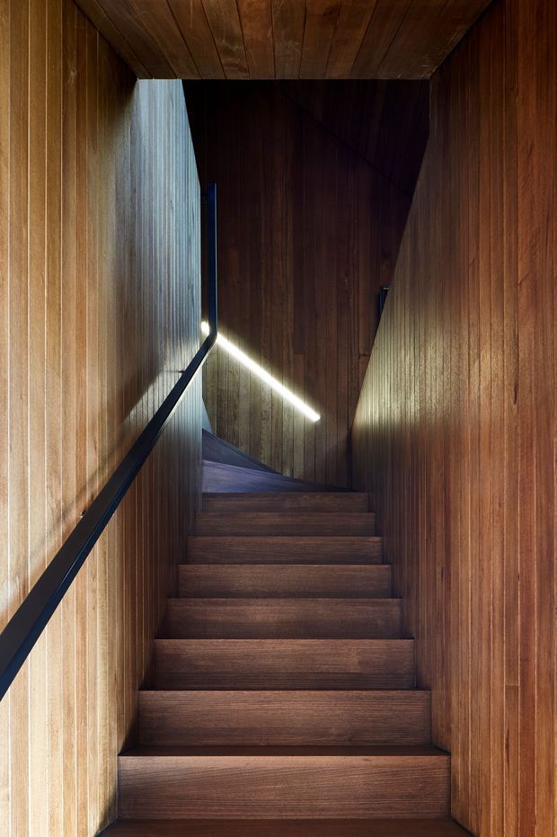 A narrow, dramatic interior stairway enclosed by richly toned vertical timber wall panelling and a matching timber-plank ceiling. Dark timber stairs rise toward a turn, with a slim black handrail and a linear integrated light creating a strong diagonal highlight. The space has a highly crafted, enclosed, contemporary architectural character.