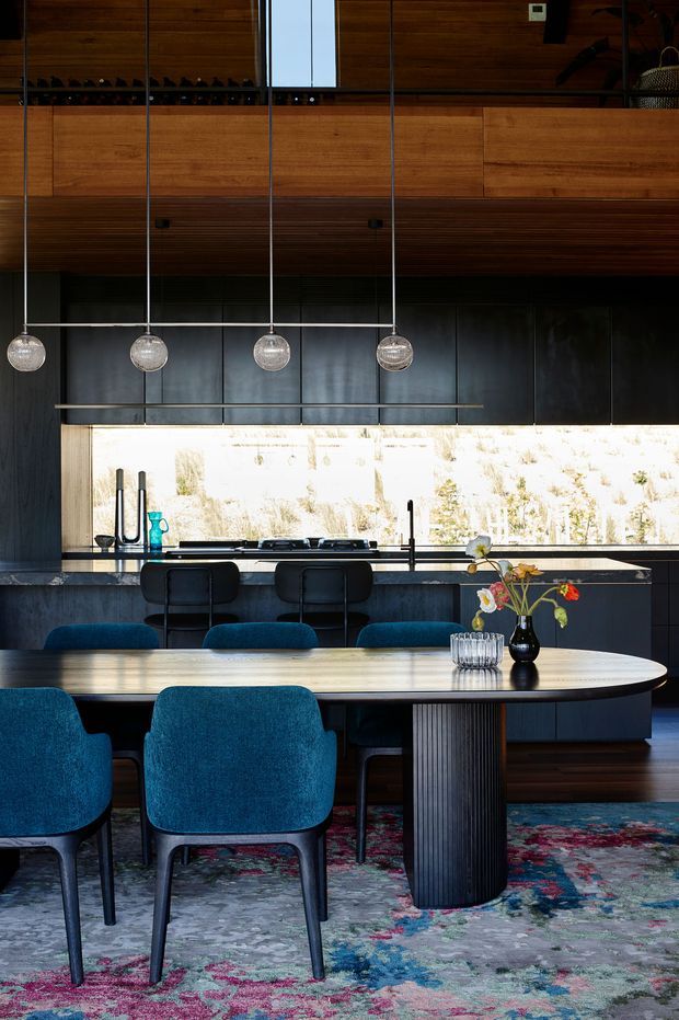 A sophisticated open-plan kitchen and dining area featuring extensive dark timber cabinetry and wall panelling, a dark stone kitchen island with seating, a dramatic horizontal window, and four spherical pendant lights. A large rounded dining table with a fluted pedestal is surrounded by rich teal upholstered chairs, set over a colourful patterned rug. The space combines warm timber, dark cabinetry, sculptural furniture, and abundant natural light.