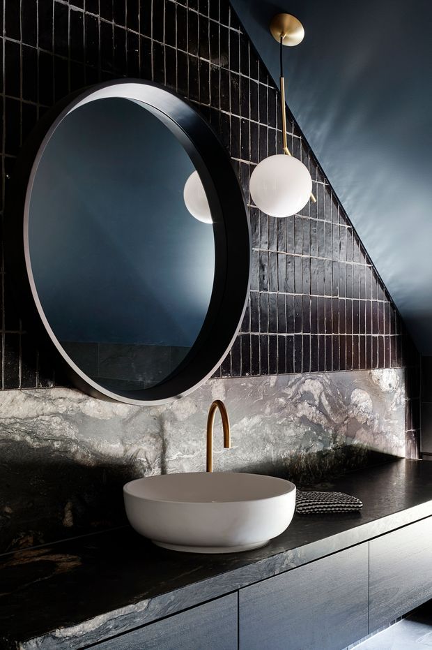 Dramatic, moody bathroom vanity with a large round black-framed mirror, black glossy tiled wall, rough natural stone backsplash, dark stone benchtop, white vessel basin, and brass wall-mounted tap. A sculptural globe pendant adds a warm metallic accent.