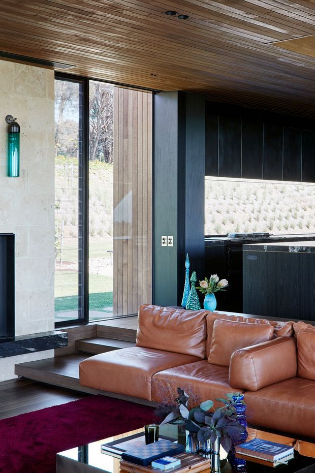 A sophisticated open-plan living room with a large cognac leather sectional sofa, dark built-in cabinetry, and a compact kitchen visible at the rear. The space features extensive timber lining across the ceiling, dark timber or metal wall panels, full-height glazing, natural stone wall cladding, stepped timber flooring, and a dramatic burgundy rug. Contemporary interiors blend warm natural materials with dark finishes and strong indoor-outdoor connection.