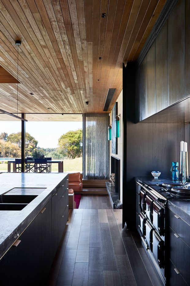 A refined contemporary kitchen with dark built-in cabinetry, a large pale stone island with integrated cooktop, and a substantial freestanding range. Warm timber lining covers the ceiling, while full-height glazing opens toward a garden and outdoor dining area. A compact lounge nook with timber flooring and a leather chair sits beyond the kitchen, creating a connected indoor-outdoor living space.