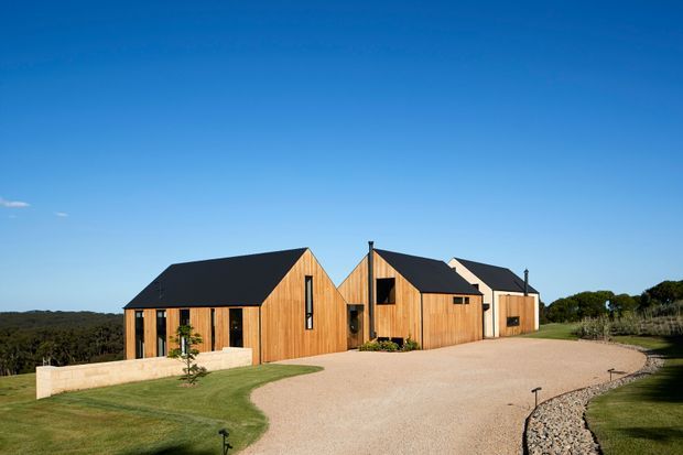 Exterior view of a contemporary multi-wing timber-clad residence set in a landscaped rural or semi-rural site. The home has steep dark gable roofs, warm vertical wood cladding, large glazed openings, and a broad pale gravel driveway curving around the front.