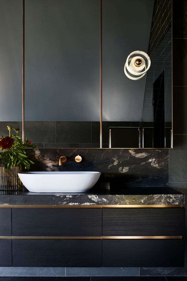 A dramatic, sophisticated bathroom vanity featuring a white vessel basin on a dark marble benchtop, black wall tiles, dark timber cabinetry, bronze-toned fixtures and mirror framing, and a sculptural wall light. The space has a moody, high-contrast palette with luxurious stone detailing and integrated storage.