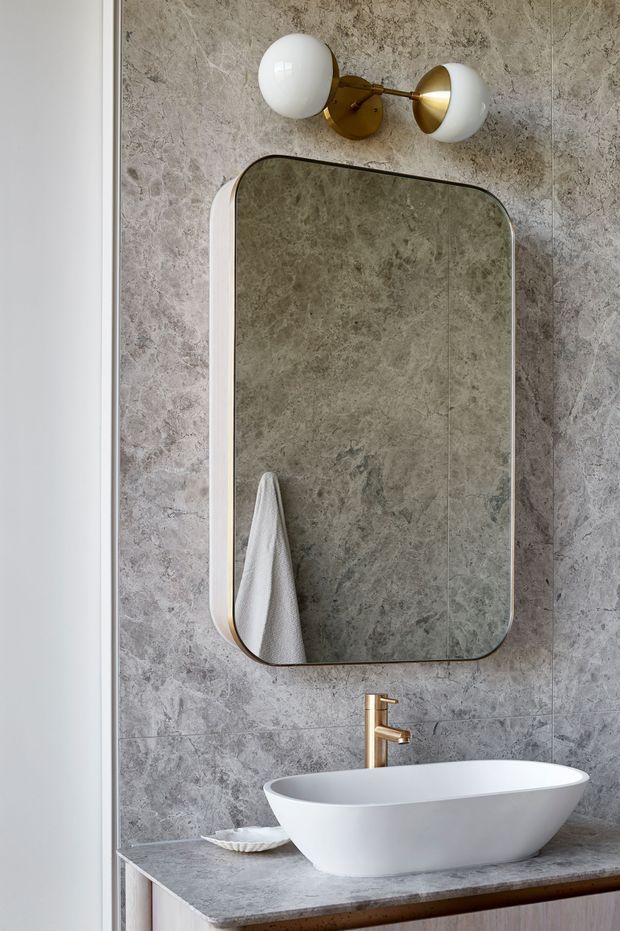 A refined bathroom vanity vignette featuring a white vessel basin on a grey stone countertop, a rounded rectangular mirror, grey veined stone wall tiles, and a brass-and-white globe wall sconce. The timber vanity edge and warm metallic fittings add contrast to the cool stone palette.