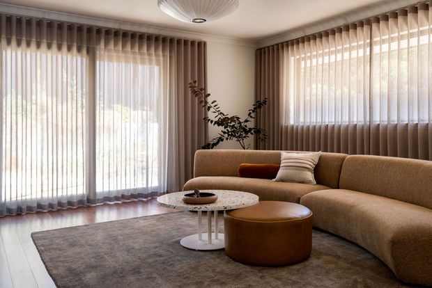 A refined living room with a large curved tan sectional sofa, round leather ottoman, marble-topped coffee table, and muted grey rug. Floor-to-ceiling windows are dressed with full-height sheer vertical curtains, creating a softly lit and private atmosphere. Neutral earthy tones, rounded furniture forms, and a sculptural ceiling light give the space a warm contemporary character.
