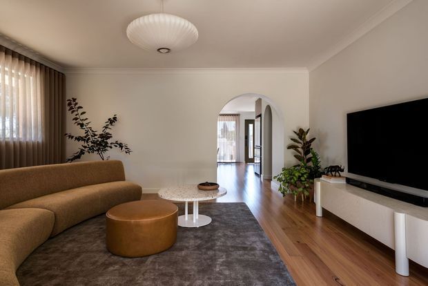 A warm, refined living room with a curved tan sofa, round leather ottoman, layered coffee tables, large textured rug, timber flooring, and a wall-mounted television on white built-in cabinetry. An arched opening leads to a bright hallway or adjoining space, while potted plants and soft layered window coverings add natural texture.