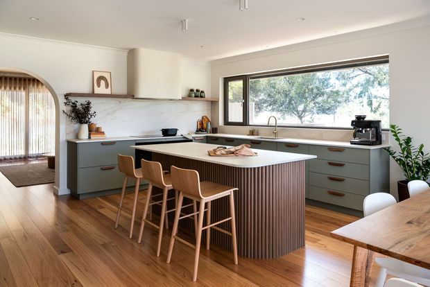 Bright, contemporary open-plan kitchen with a large island featuring vertical fluted timber detailing, muted blue-grey cabinetry, a pale stone benchtop and warm timber flooring. Three timber bar stools sit at the island, with a timber dining table partly visible in the foreground. A wide framed window overlooks trees, while an arched opening connects to an adjacent space.