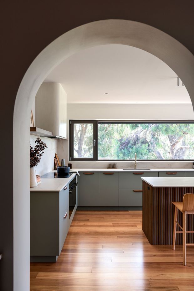 A bright contemporary kitchen viewed through a large rounded archway. The room features muted green-grey cabinetry with warm timber handles, white stone benchtops, a timber floor, dark-framed horizontal windows, and a central island with a vertical timber-slatted base and bar seating. Natural light and greenery are visible through the windows.