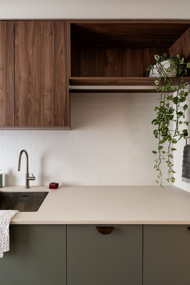 Close-up view of a contemporary kitchen with a warm walnut overhead cabinet and open shelf, matte muted green base cabinetry, a pale beige engineered-stone countertop, white subway-tile splashback, and a dark undermount sink with brushed metal gooseneck tap. A trailing potted plant adds organic detail to the open shelving.